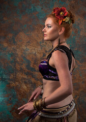 Fototapeta na wymiar side view of beautiful woman in belly dance costume with black lace in red circlet of flowers looking away and holding hands near in tribal pose in front of teal and orange background