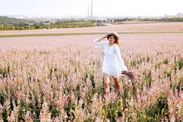 Fototapeta na wymiar Young brunette woman wearing a straw hat and a white dress, running along a field with pink sage flowers at sunset. Beautiful girl relaxing outdoors, having fun, holding plant.