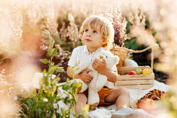Little blonde boy wearing a straw hat, sitting in a field at sunset, feeding a plush bear toy a...