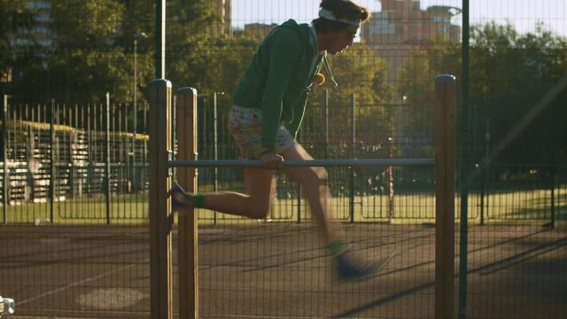 Funny freak man in green hoodie, color shorts and sunglasses pushing up on parallel bars and actively waving legs as if he was running somewhere. Sport humor concept.