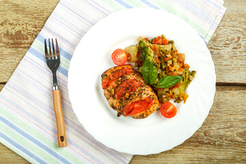 Baked chicken breast with tomatoes with a side dish of vegetables on a white plate with a fork on a linen napkin.