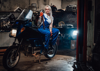 Obraz na płótnie Canvas Tattooed hipster girl in work overalls hold big wrench while sitting on sportbike in garage or workshop, smiling and looking on camera