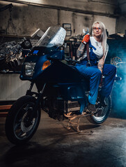 Fototapeta na wymiar Young blond woman mechanic in work overalls hold big wrench while sitting on sportbike in garage or workshop, smiling and looking on camera