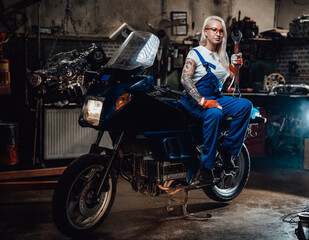 Obraz na płótnie Canvas Young blond woman mechanic in work overalls hold big wrench while sitting on sportbike in garage or workshop