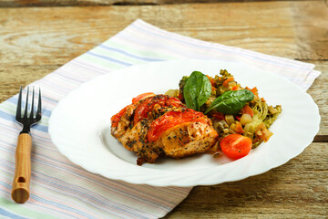 Baked chicken breast in spices with tomatoes with a side dish of vegetables on a white plate with a fork on a napkin.