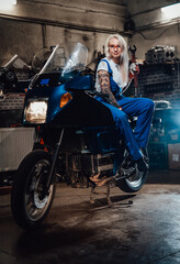 Plakat Tattooed hipster girl in work overalls hold big wrench while sitting on sportbike in garage or workshop, smiling and looking on camera