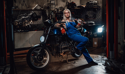 Fototapeta na wymiar Tattooed hipster girl in work overalls hold a big wrench and posing for a camera while leaning on her naked bike in garage or workshop