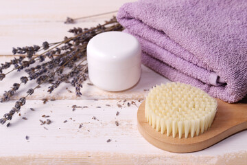 Fototapeta na wymiar Massage wooden brush with natural bristles, a white jar of cream, a fresh lilac Terry towel neatly folded and a bouquet of dry lavender on a white wooden background. Spa procedures. Selective focus