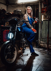Obraz na płótnie Canvas Tattooed hipster girl in work overalls hold a big wrench while leaning on her naked bike in garage or workshop, smiling and looking on camera