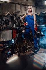 Fototapeta na wymiar Tattooed hipster girl in work overalls hold a big wrench and posing for a camera while standing on naked bike in garage or workshop