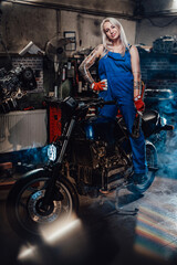 Fototapeta na wymiar Tattooed hipster girl in work overalls hold a big wrench and posing for a camera while standing on naked bike in garage or workshop