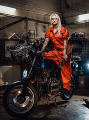 Obraz na płótnie Canvas Tattooed hipster girl in orange overalls hold a big wrench and posing for a camera while standing on her naked bike in garage or workshop, smiling and looking on camera