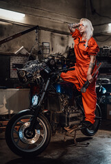 Fototapeta na wymiar Tattooed hipster girl in orange overalls hold a big wrench and posing for a camera while standing on her naked bike in garage or workshop, smiling and looking on camera
