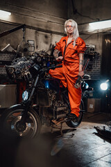 Fototapeta na wymiar Tattooed hipster girl in orange overalls hold a big wrench and posing for a camera while standing on her naked bike in garage or workshop