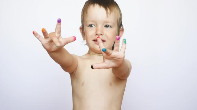 A cute little boy stands with arms extended forward showing his painted multi-colored fingers against white background
