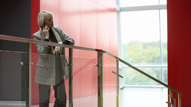A side view panoramic shot of a middle-aged female executive standing inside the office centre near the staircase and recieving a call on her phone from her business partner