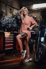 Fototapeta na wymiar Charming blonde woman wearing pink dress posing in garage or workshop, standing next to suspended flat engine and naked bike