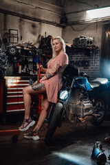 Fototapeta na wymiar Beautiful blonde girl with tattooed body wearing pink dress posing next to the suspended flat engine and naked bike in garage or workshop