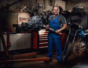 Fototapeta na wymiar Professional mechanic stands next to the suspended flat engine and naked bike in garage or workshop, smiling and looking on camera
