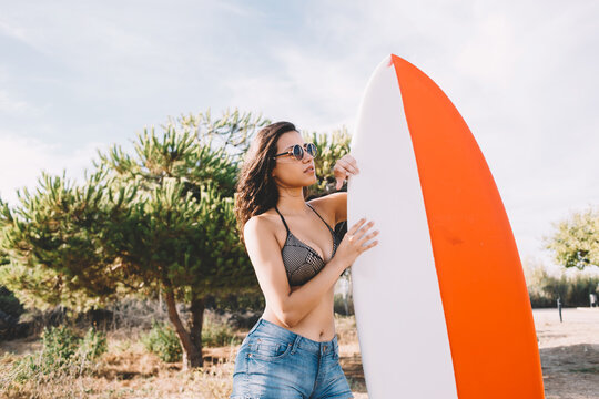Half length portrait of an attractive brunette hair model posing with her surfboard in the fresh air in sunny day, young alluring women relaxing after surfing in the ocean during summer weekend