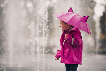 Happy child girl 4-5 year old holding pink umbrella wearing raincoat over rain at background...