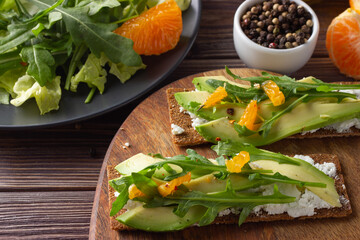 Wholegrain crisp bread with cheese, fresh rocket leaves, avocado and tangerine with green salad on wooden background. Close up.