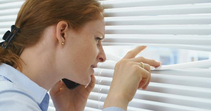 Stressed business woman looking out through window blinds and reporting an accident on the phone.