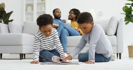 Happy african american family spending time together at home. Parents relax on sofa in living room together, small children brother and sister reading book at home, family time concept
