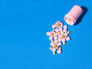 On a blue background, white and yellow tablets spilling out of a jar with chamomile flowers. The concept of natural medicines