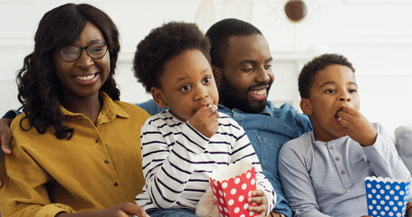 Happy african american family watching tv, eating popcorn snack, having fun at home.