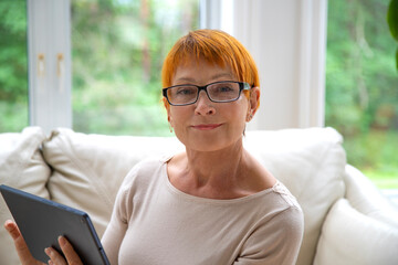 Portrait of mature, Caucasian woman wearing eyeglasses using electronic tablet at home.