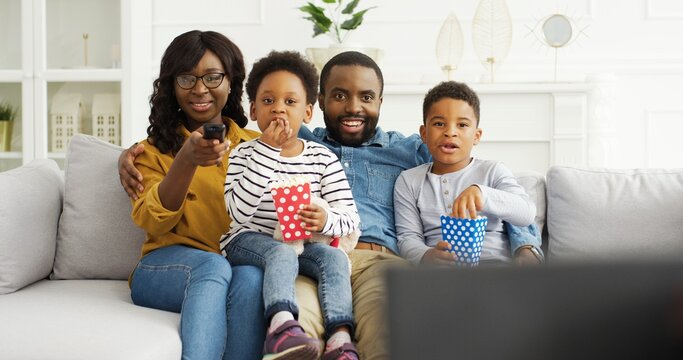 African American Parents With Little Kids Watching Funny Movies Or Tv Show, Eating Popcorn, Resting On Sofa At Home.