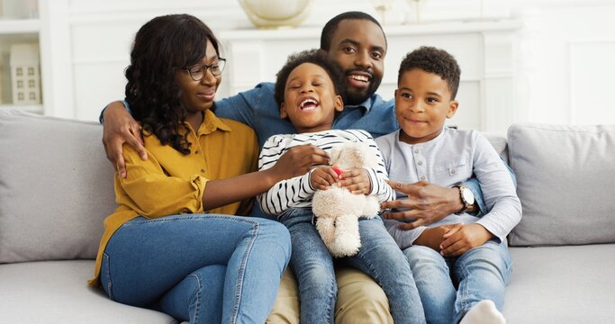 Portrait Of African American Parents With Little Kids Resting On Sofa At Home. Family Celebrating 4th Of July, Patriotic Holiday Day.