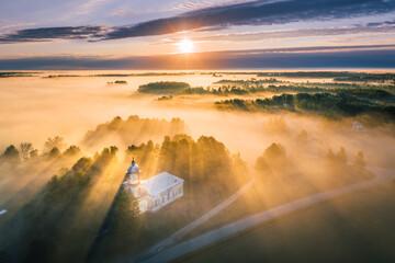 Majestic church covered in fog and surrounded by forest. Aerial view over picturesque countryside with at sunrise with long light rays. 