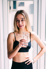 Young smiling woman in sporty top and leggings sitting drinking pure water while joyfully looking in camera at home with big beautiful windows