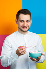 A man on a yellow, blue, orange, pink background in a white shirt with a turquoise plate of marshmallows and a pink spoon. The guy with a smile. An image for a male photo. Photo session concept