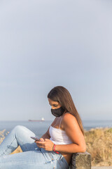 young woman with mask looks at her phone sitting on a bench overlooking the sea