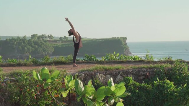 Handheld shot of man in tank top and leggings doing upward salute and bending backwards in yoga pose while practicing on cliff by ocean