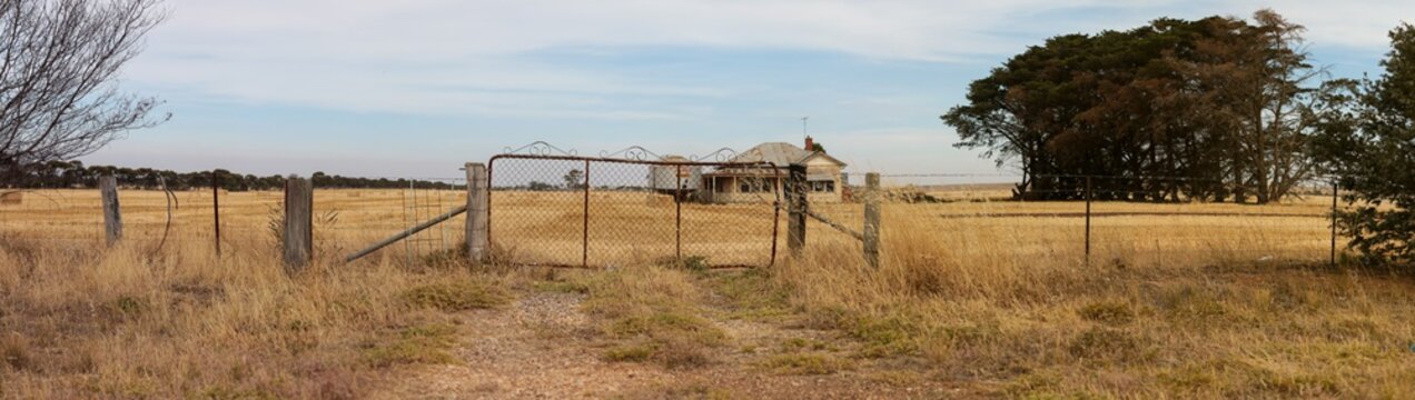 Panoramic image of an old farm gate and a timber worn out abandoned traditional Australian farm house in the middle of a newly harvested field on a agricultural property in rural Victoria, Australia