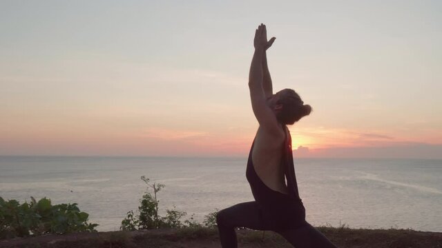 Handheld dolly-out shot of male yogi in tank top and leggings standing on mat on cliff by ocean and doing warrior pose at sunset