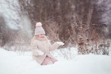 Fototapeta na wymiar Cute little girl child blonde in a pink knitted hat with a pompom and a fur coat sitting in a snowdrift in the winter outdoors throwing snow. Winter walks, fun outdoors winter time.