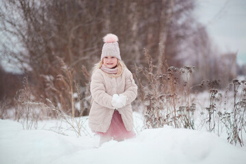 Cute baby blonde girl in a pink knitted hat with a pompom and a fur coat standing outdoors in winter holding snow in her hands. Winter time