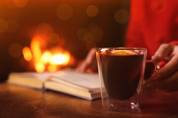 Woman with mulled wine reading book near fireplace indoors, closeup. Winter vacation