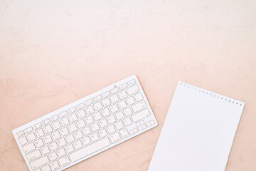 Pastel pink desktop and white keyboard wireless on a wooden surface, blank notepad sheet