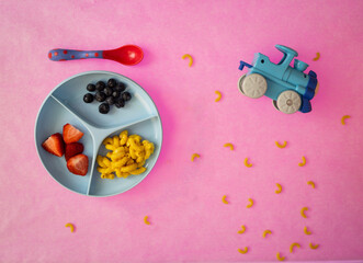 Toddler plate with macaroni and cheese, strawberries and blueberries with toy train around the food on pink background with pasta spread around
