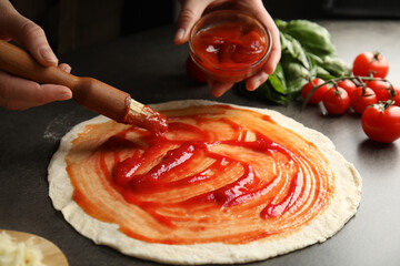 Woman spreading tomato sauce onto pizza crust at grey table, closeup