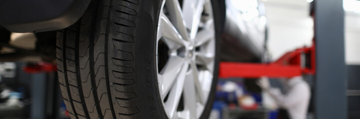 Close-up view of car all-season tire with aluminum rim. Mechanical lift for vehicle. Automobile service center or workshop concept. Professional auto maintenance - obrazy, fototapety, plakaty