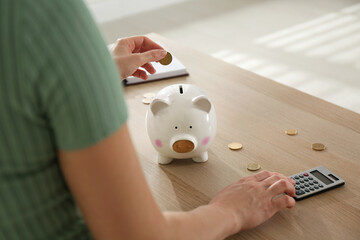 Woman putting money into piggy bank and using calculator at wooden table indoors, closeup