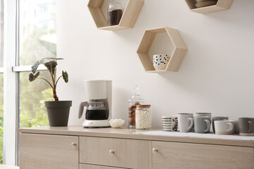 Modern coffeemaker and cups on commode near light wall indoors
