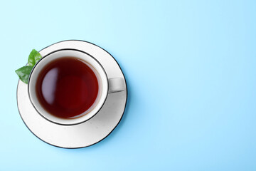 Cup of aromatic black tea and green leaves on light blue background, top view. Space for text